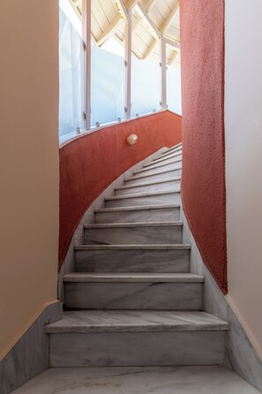A marble staircase with a red wall on one side and a white wall on the other, leading to a room with a sloped ceiling and large windows. - Red Tower (Lefkada)