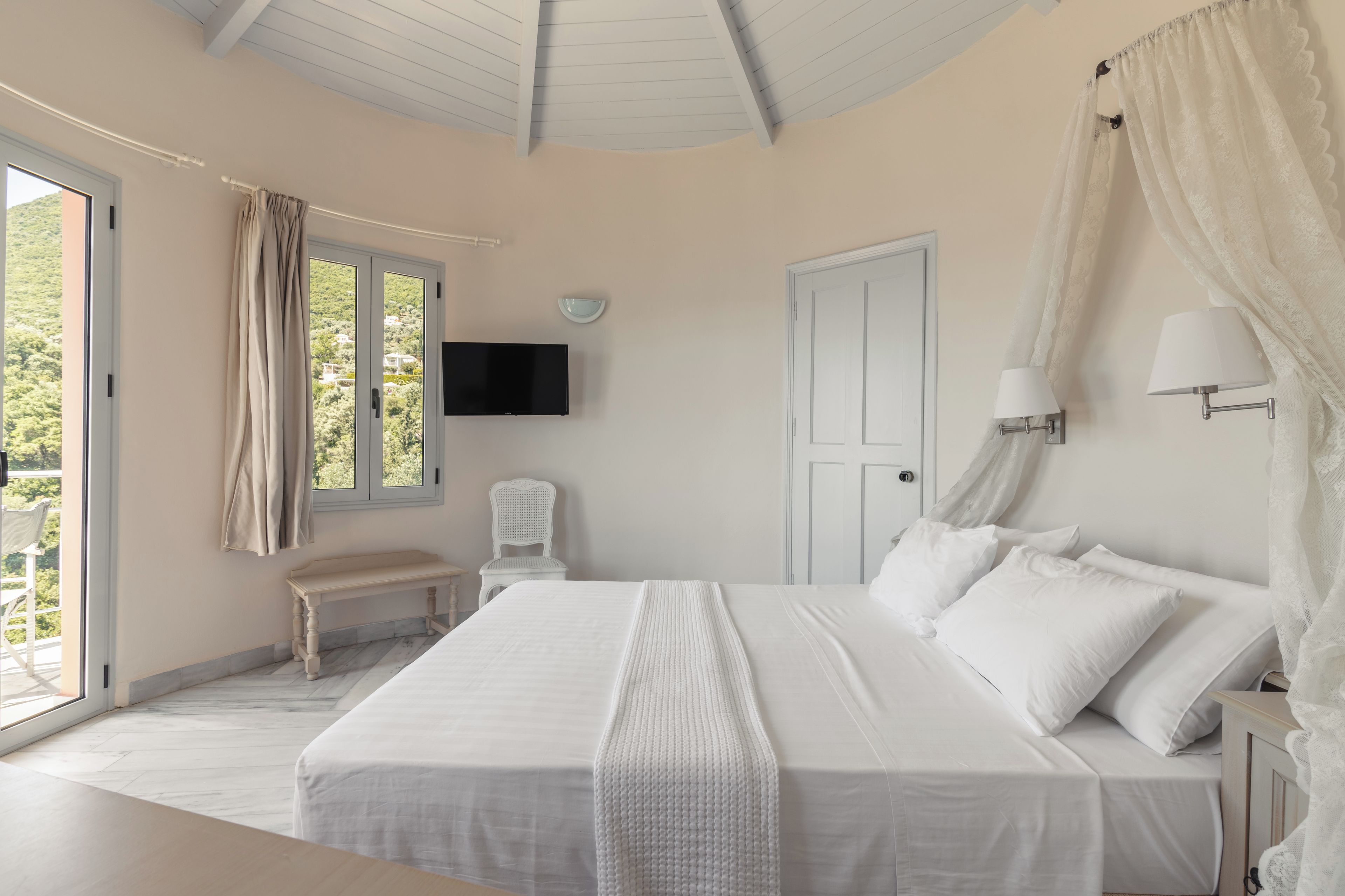 Quarto com uma cama grande, um banco, uma televisão e vista para o verde lá fora.