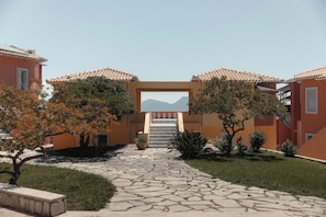 A courtyard with a stone pathway leading to a staircase, surrounded by buildings with terracotta roofs and a mountain view. - Red Tower (Lefkada)