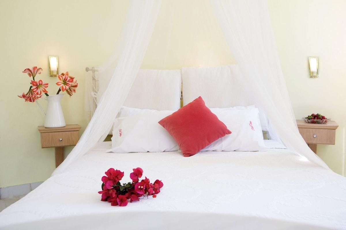 Una cama con ropa blanca, un almohadón rojo y un ramo de flores rojas.