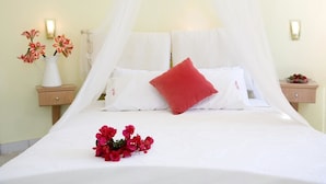 Una cama bien hecha con sábanas blancas, un cojín rojo y un ramo de flores rojas. Hay dos mesitas de noche con lámparas y un florero con flores en una de ellas.