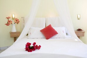 Una cama bien hecha con sábanas blancas, un cojín rojo y un ramo de flores rojas. Hay dos mesitas de noche con lámparas y un florero con flores en una de ellas.