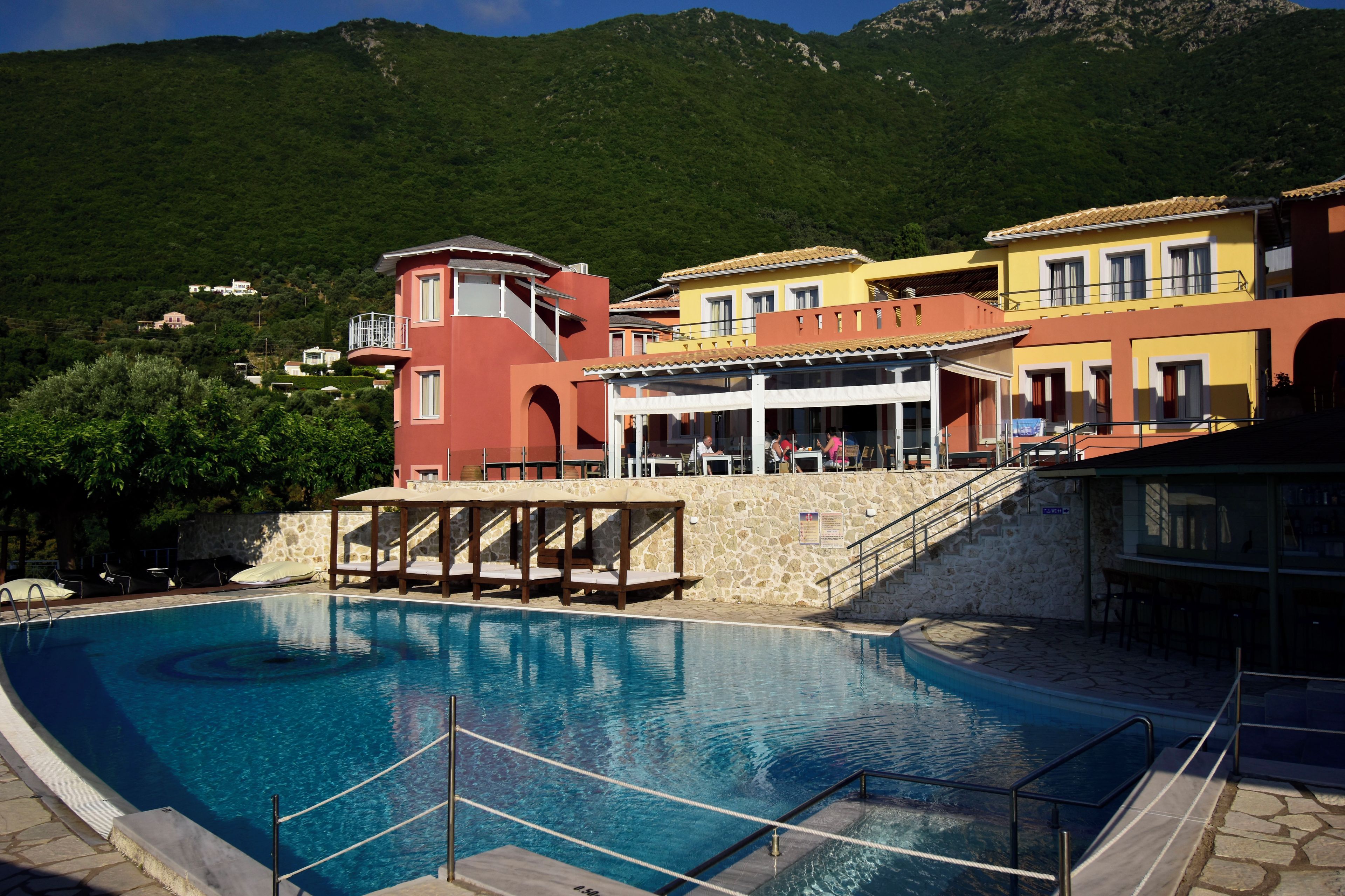 A swimming pool with a stone deck, surrounded by colorful buildings with balconies and a mountain backdrop.