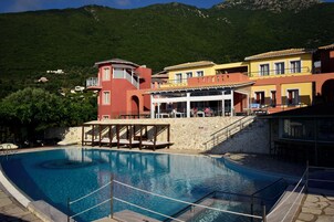 Una piscina con solárium de piedra, rodeada de edificios coloridos con balcones y montañas al fondo.