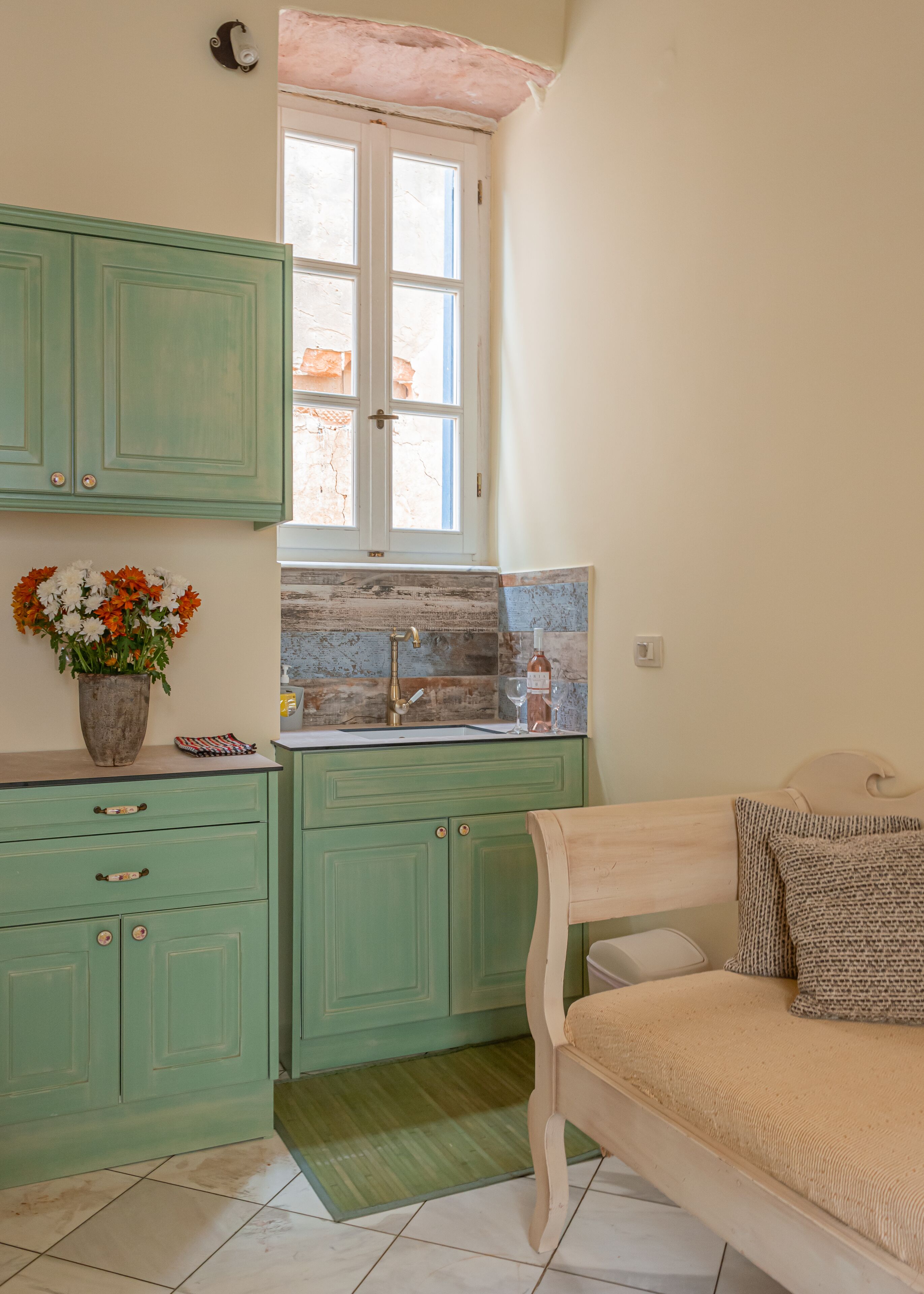 A small, cozy kitchen with green cabinets, a window, and a bed with a beige blanket.
