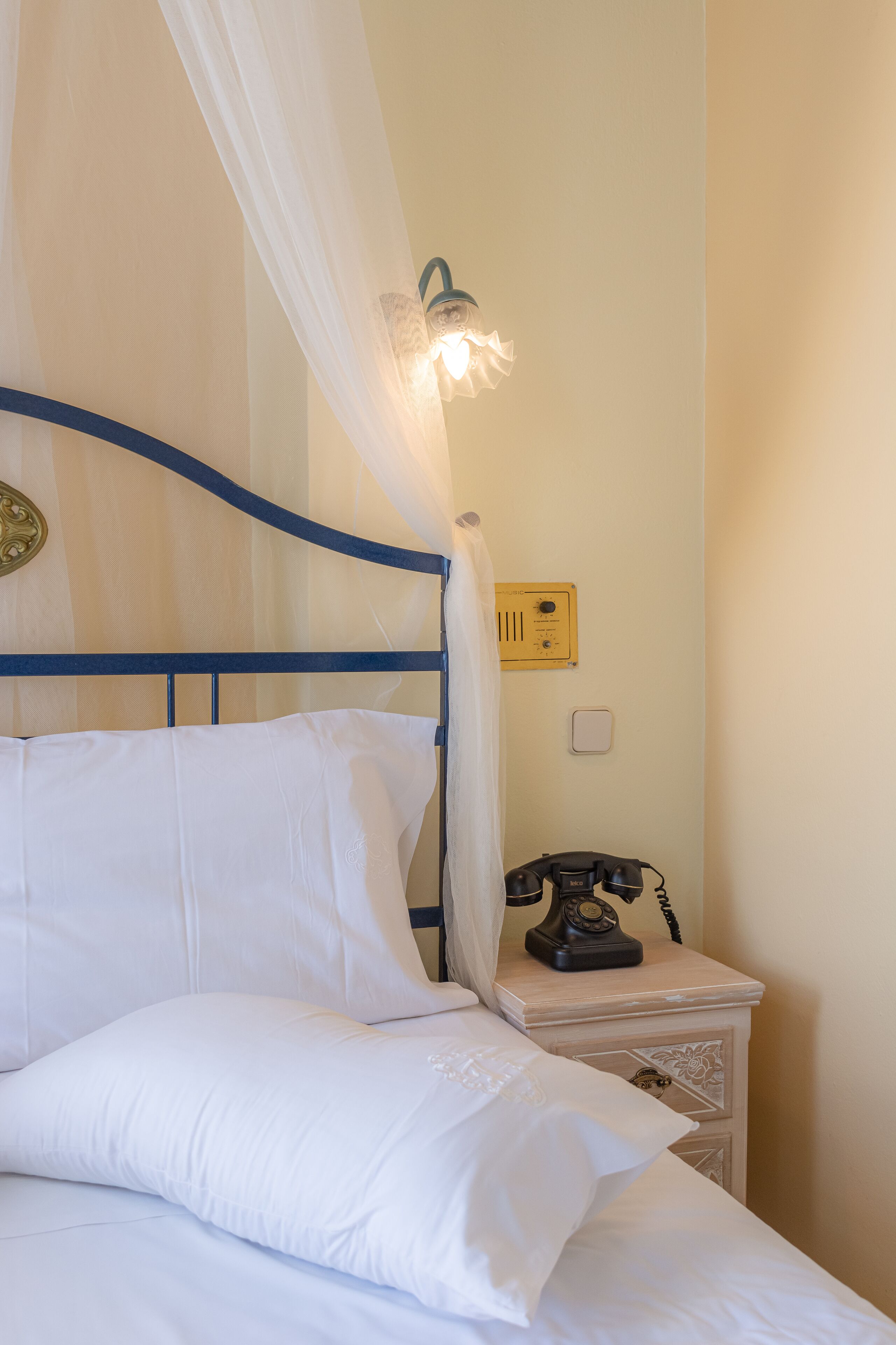 A bed with white linens, a blue metal headboard, and a white canopy. There is a bedside table with a rotary phone and a wall-mounted light fixture.