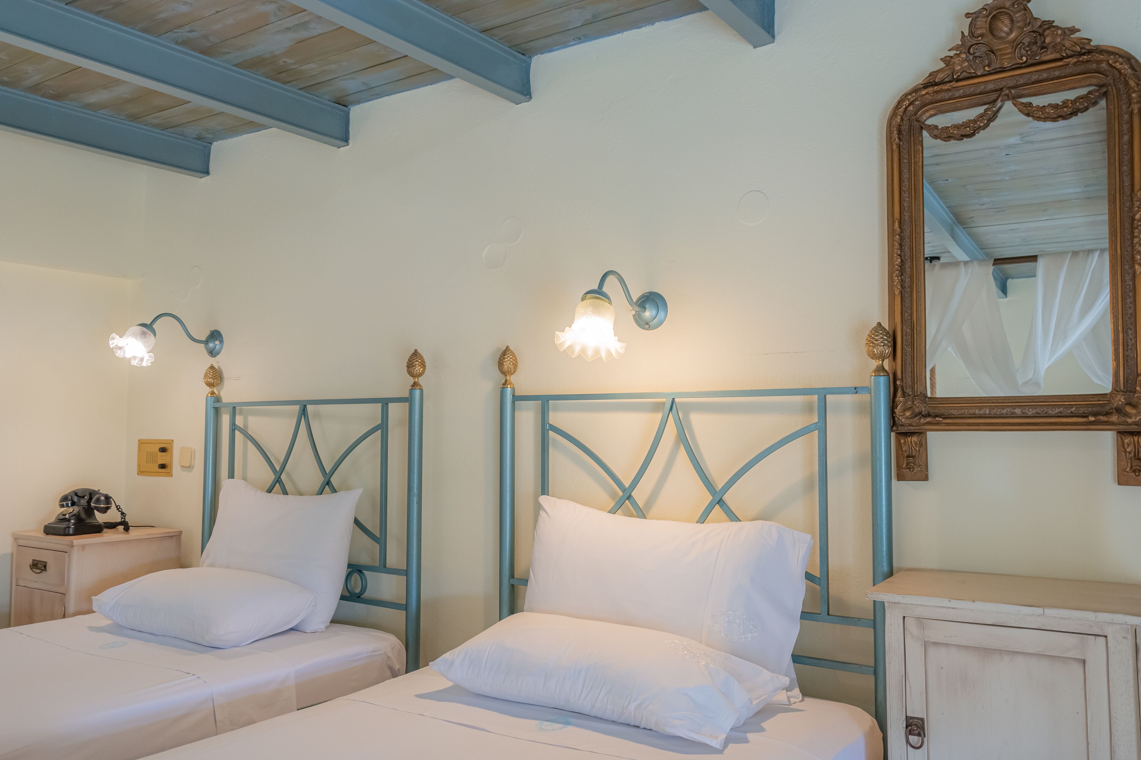 A room with two beds, each with a blue headboard and white bedding. There are wall sconces above the beds, a wooden cabinet, and a large mirror on the wall.