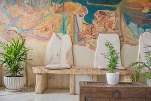 A stone bench with white cushions, two potted plants, and a wooden chest in a room with a colorful mural on the wall.