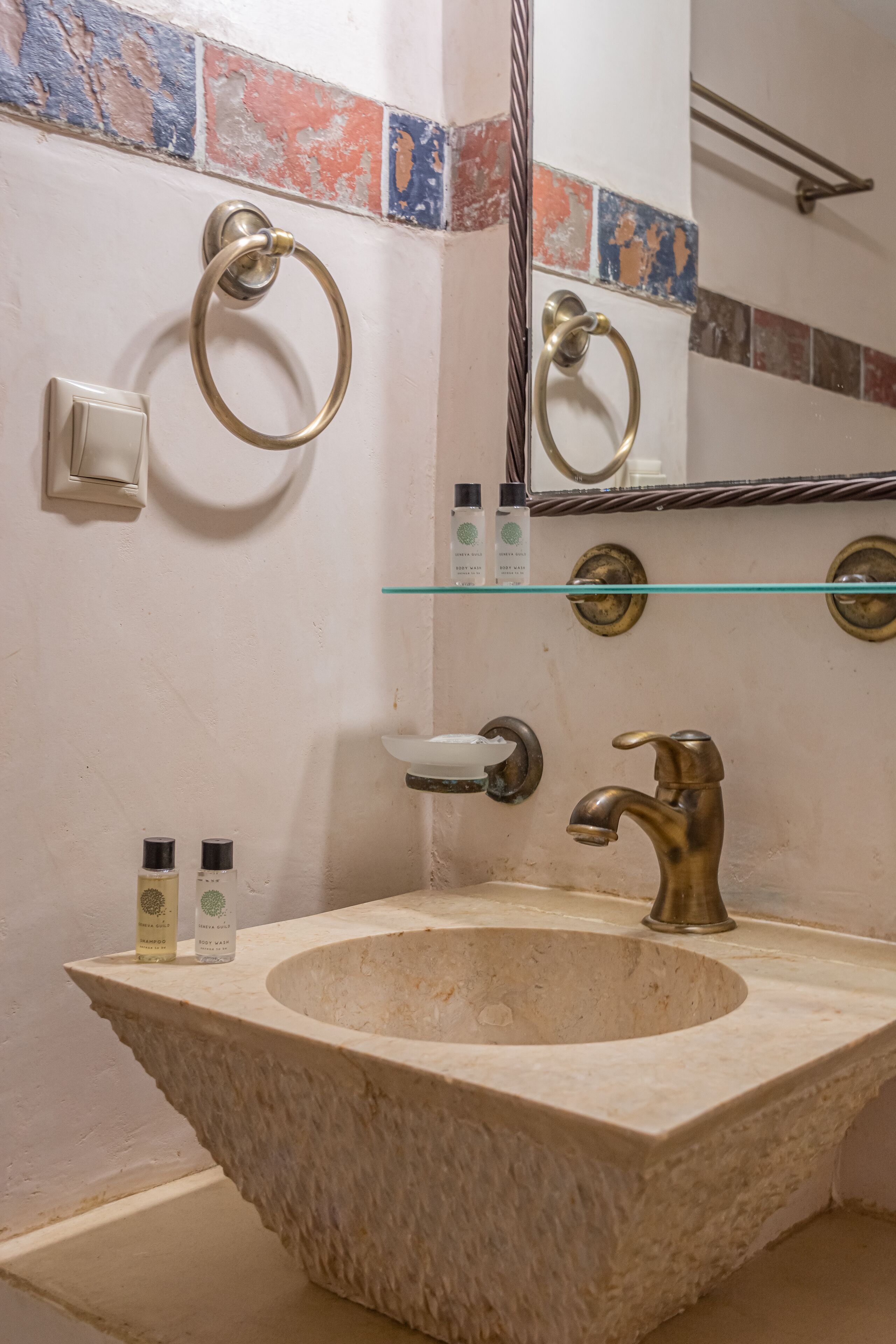 A bathroom with a stone sink, brass fixtures, and a glass shelf.