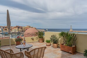 A terrace with wicker chairs, a table, potted plants, and a view of the sea and buildings. - Pandora Suites (Chania)