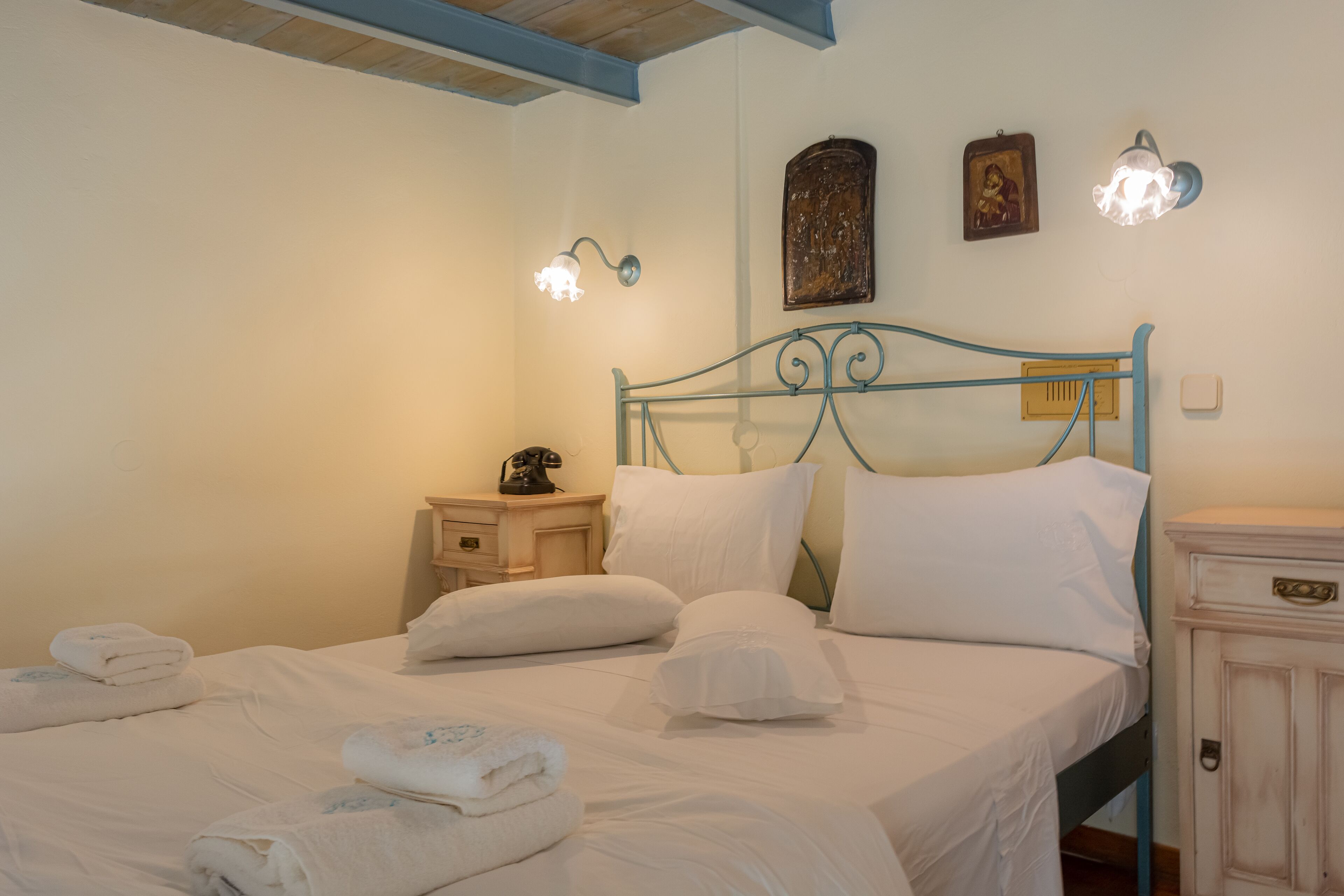 A neatly made bed with white linens and folded towels, a bedside table with a lamp, and a small wooden cabinet.