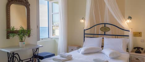 A neatly arranged bedroom with a bed, bedside tables, a mirror, and a view of the sea through a window.