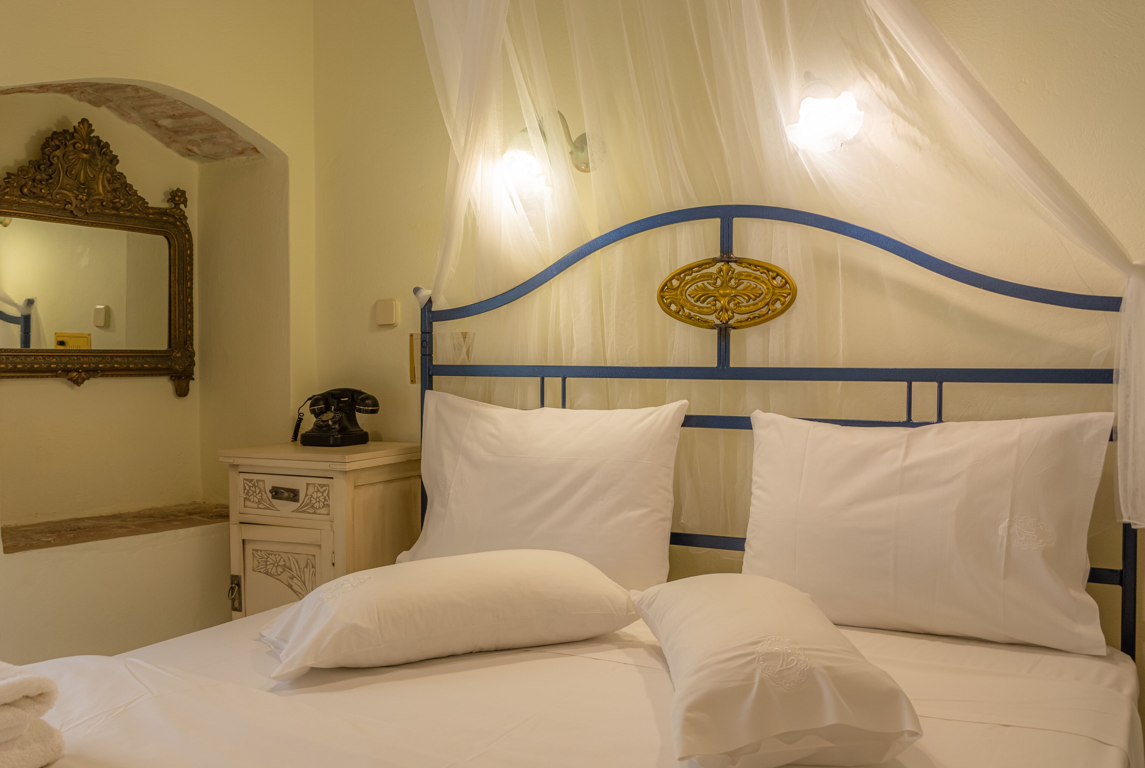 A neatly made bed with white linens and pillows, a bedside table with a telephone, and a decorative mirror on the wall.