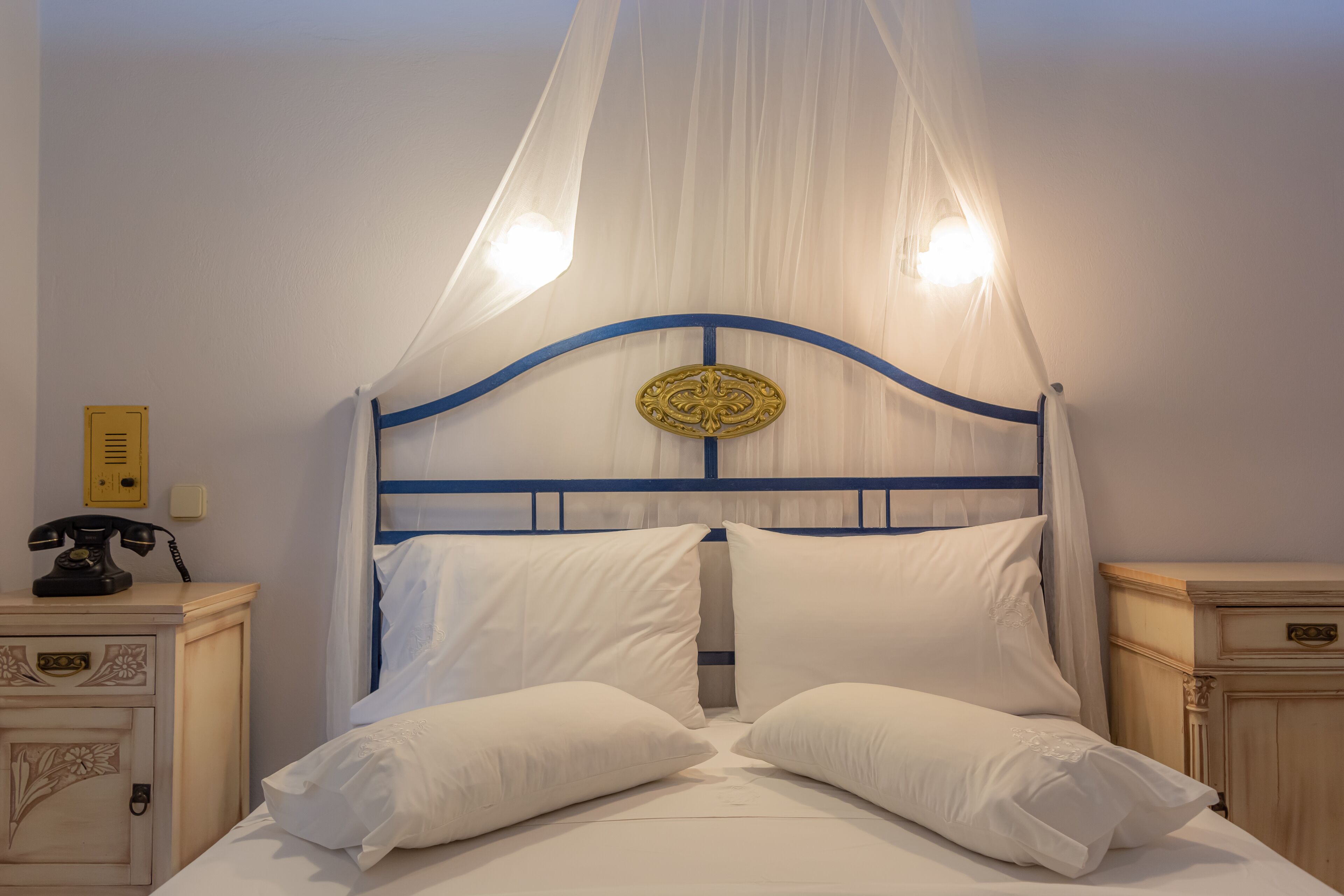 Un lit avec du linge de lit blanc et un baldaquin, un téléphone sur la table de chevet et une petite table de nuit avec un meuble décoratif.