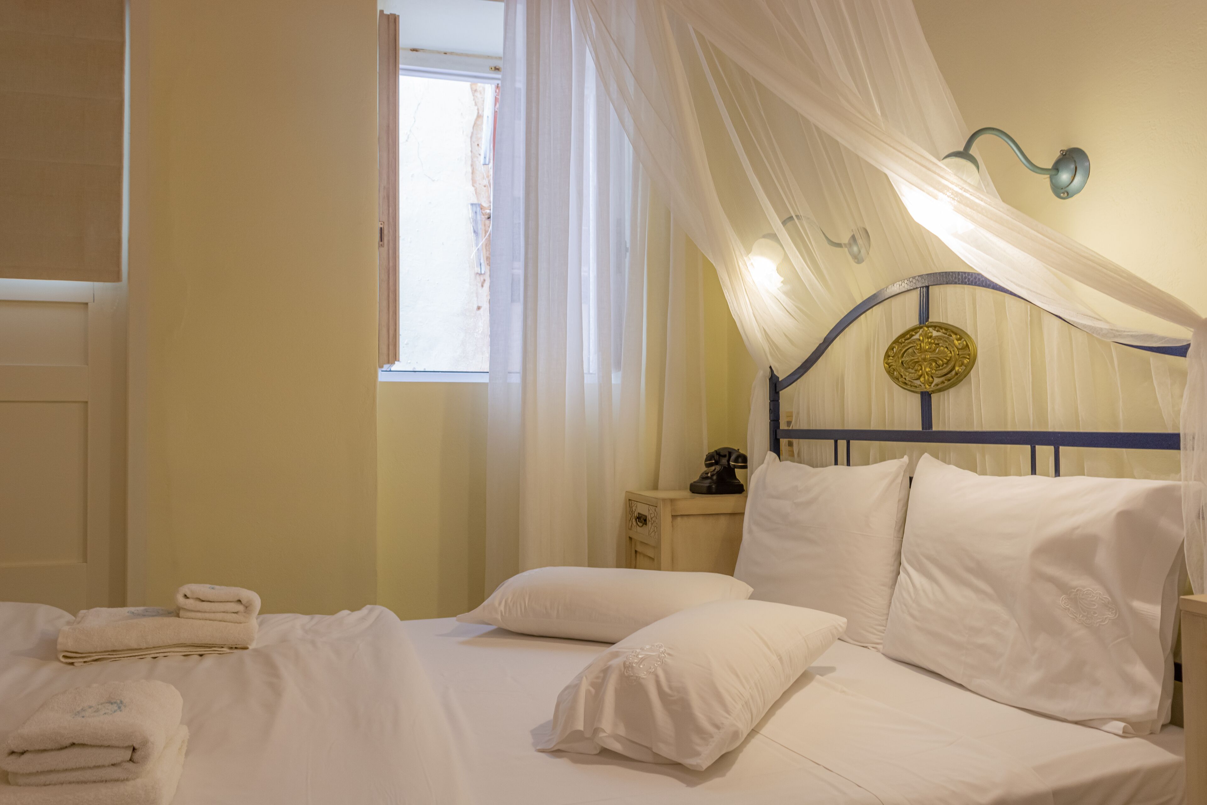 A neatly made bed with white linens, a bedside table with a lamp, and a window with sheer curtains.