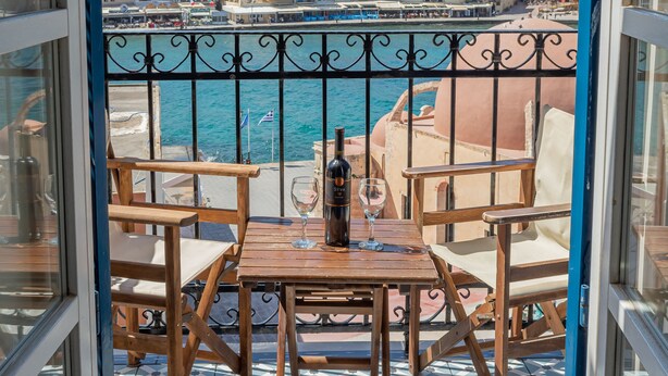 A balcony with a table and chairs, overlooking a coastal town with buildings and a clear sky. - Pandora Suites (Chania)