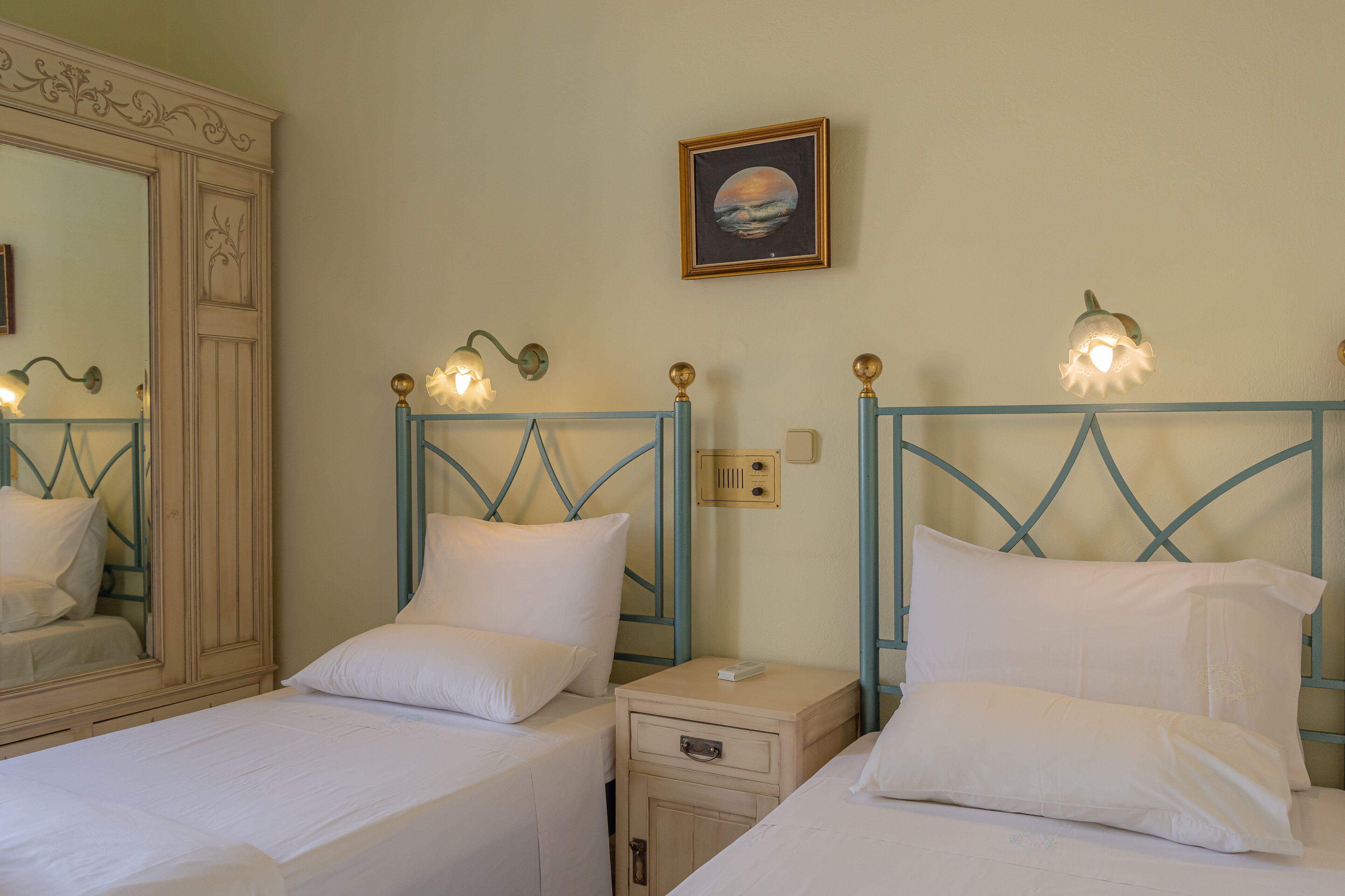 Une chambre d’hôtel avec deux lits, chacun doté de draps blancs et d’une tête de lit en métal bleue. Il y a une table de chevet avec une lampe et un petit tiroir. Un tableau encadré représentant un coucher de soleil est accroché au mur au-dessus des lits.