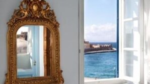 A room with a large ornate mirror on the wall, a window with a view of the sea, and a white door. - Pandora Suites (Chania)