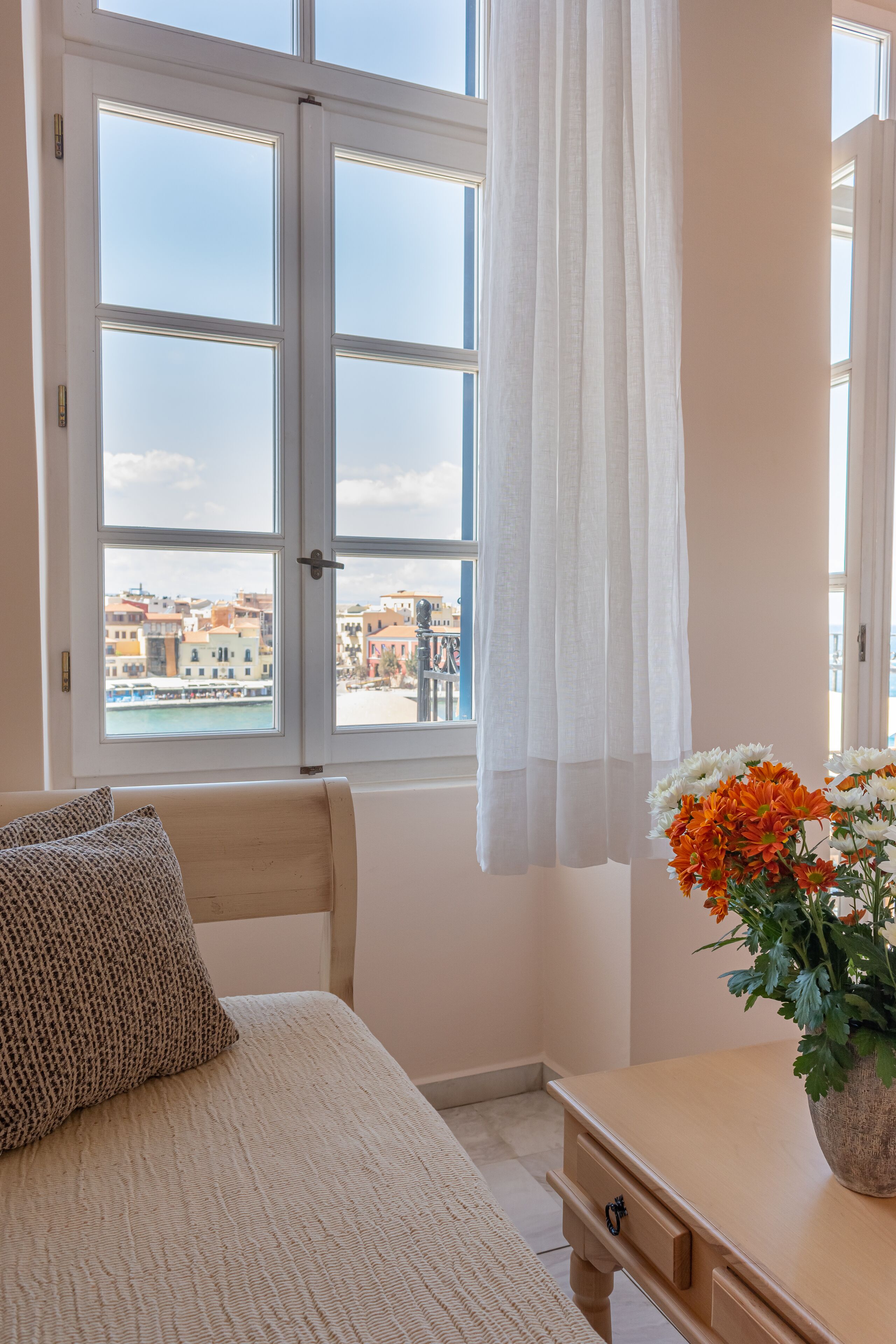 A bedroom with a bed, a nightstand, a window with curtains, and a view of buildings and water.
