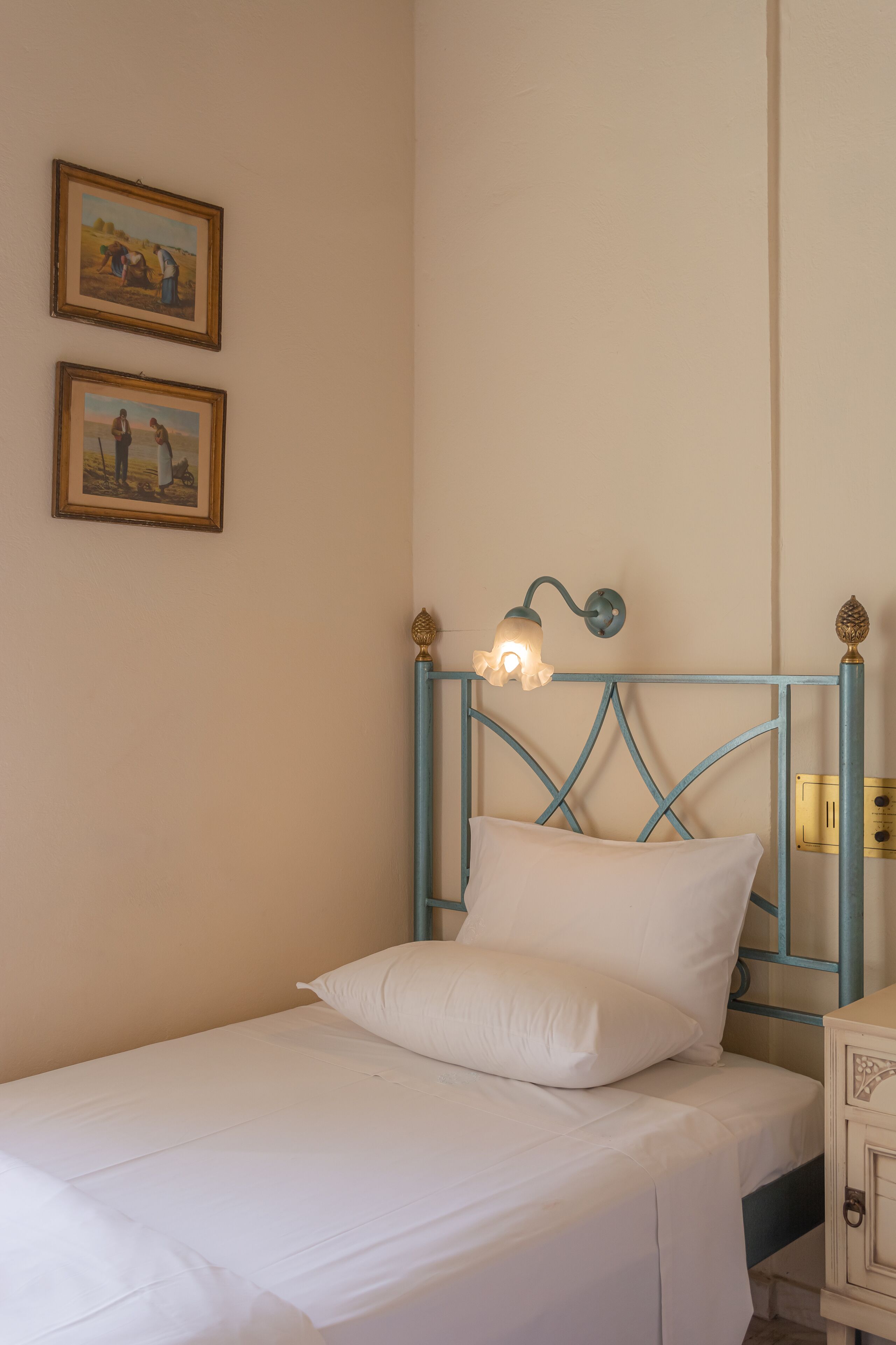 Una cama individual con colchón y almohadas blancos, cabecero de metal azul y dos cuadros colgados en la pared.