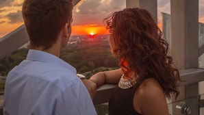 Una pareja contempla la puesta de sol desde un balcón con vista a la ciudad.