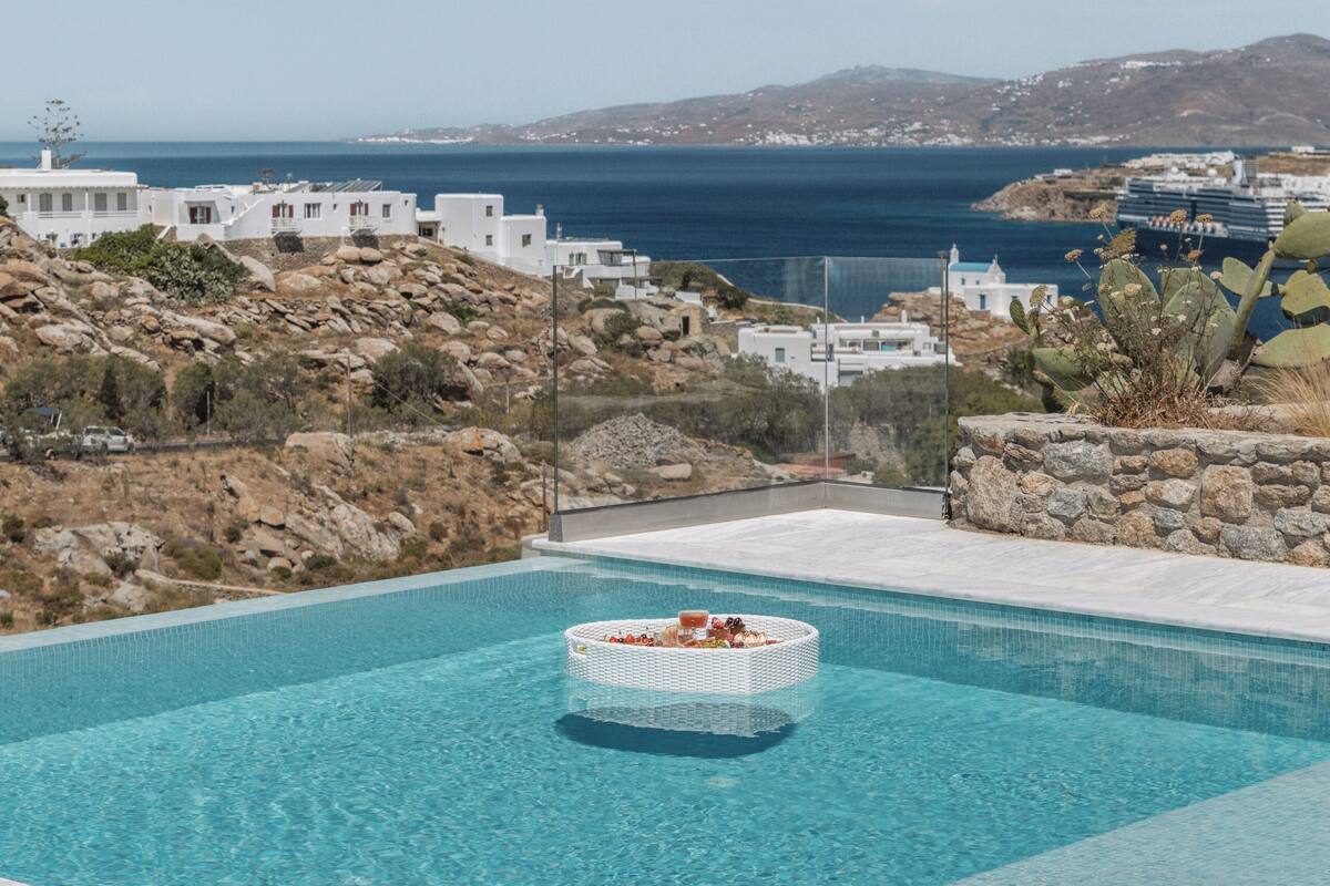 En pool med ett avslappningsområde, utsikt över havet och vita byggnader i bakgrunden.