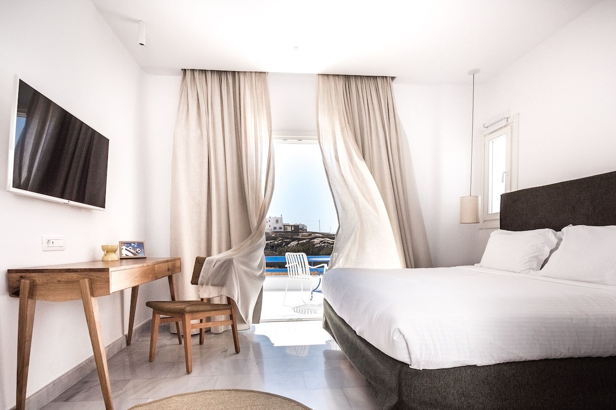 Ett modernt hotellrum med en säng, ett skrivbord, en stol och utsikt över havet.