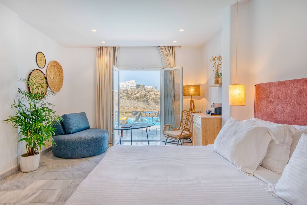 Ett modernt hotellrum med en säng, en sittgrupp med en blå fåtölj, ett litet bord och en balkong med utsikt över byggnader och en pool.
