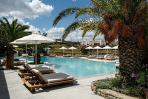 Un espace piscine agrémenté de chaises longues et de parasols, entouré de palmiers et surplombé d’un ciel dégagé.