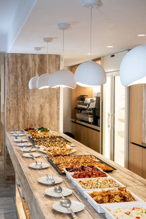 A buffet with various dishes, a marble wall, and hanging pendant lights. - San Antonio Summerland (Mykonos)