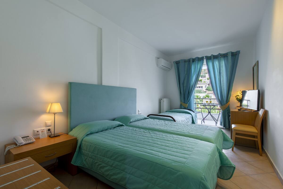 A hotel room with two beds, a desk with a lamp, a chair, and a view of a balcony with a table and chairs.