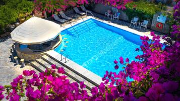 Una piscina con sillas de descanso y un área de estar cubierta, rodeada de coloridas flores de buganvilla.