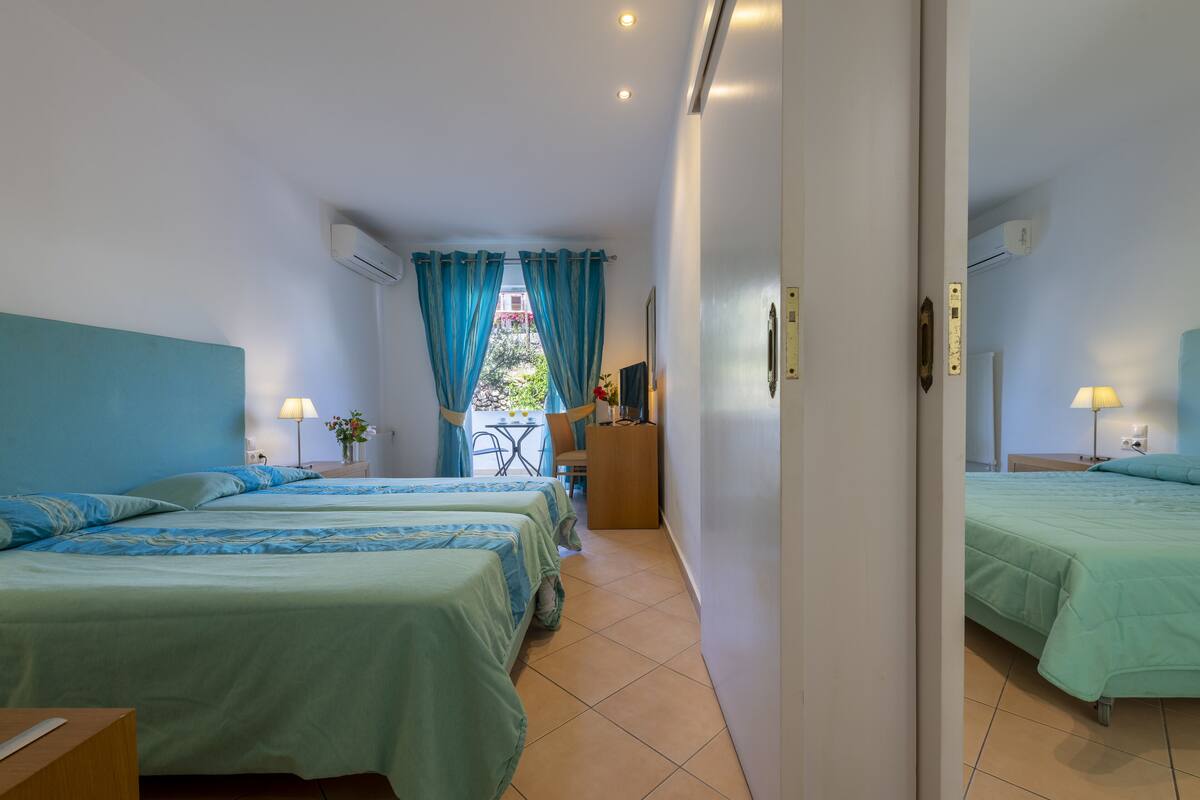 A hotel room with two beds, a desk, a chair, and a view of the outdoors through a window with blue curtains.