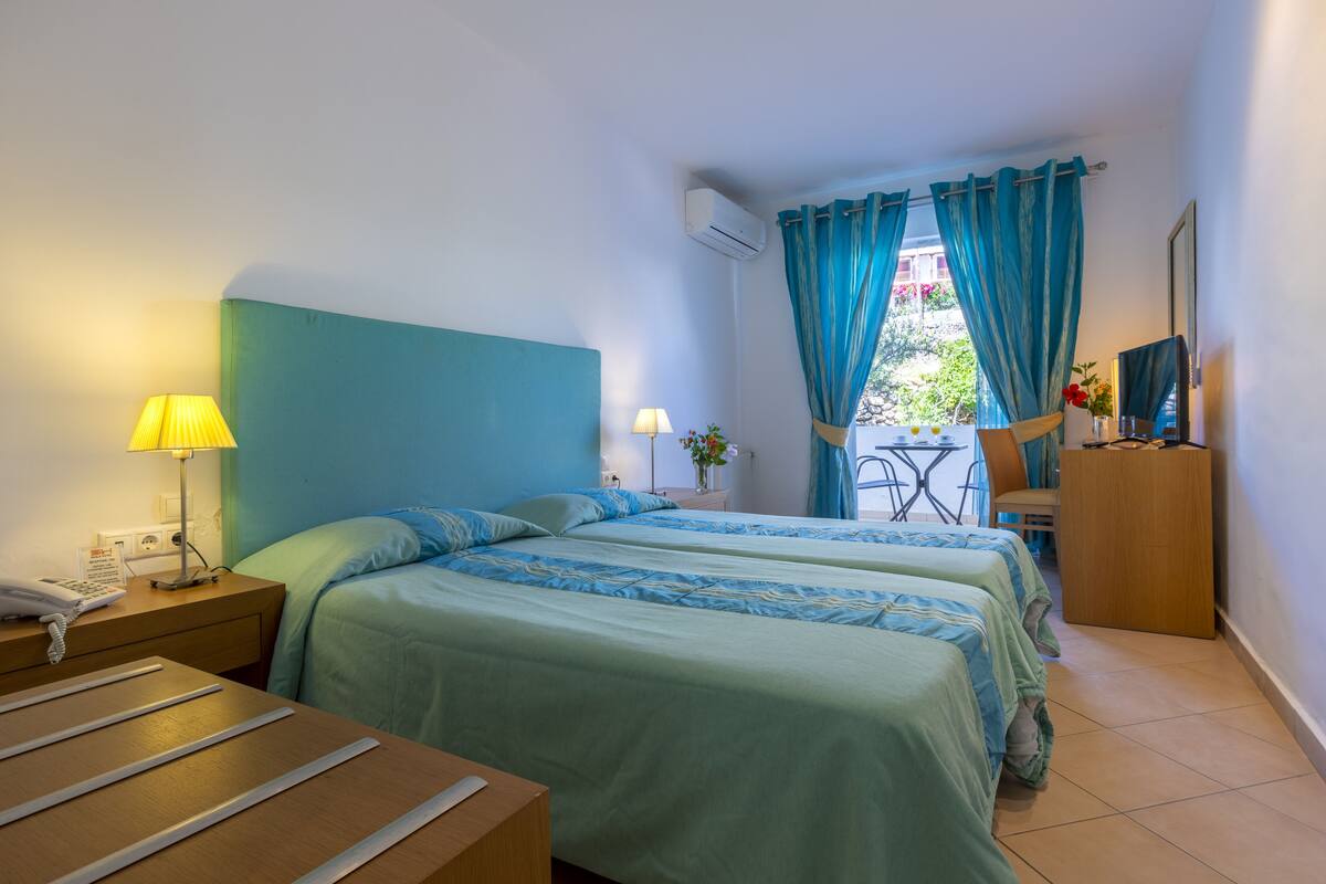 A hotel room with two beds, a desk, a chair, a small table, a lamp, an air conditioner, and a window with curtains.