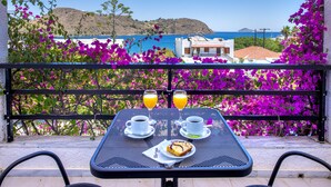 Une table sur un balcon, avec deux tasses de café, une assiette de nourriture et deux verres de jus d’orange, offrant une vue sur la côte, avec des fleurs violettes et des bâtiments.