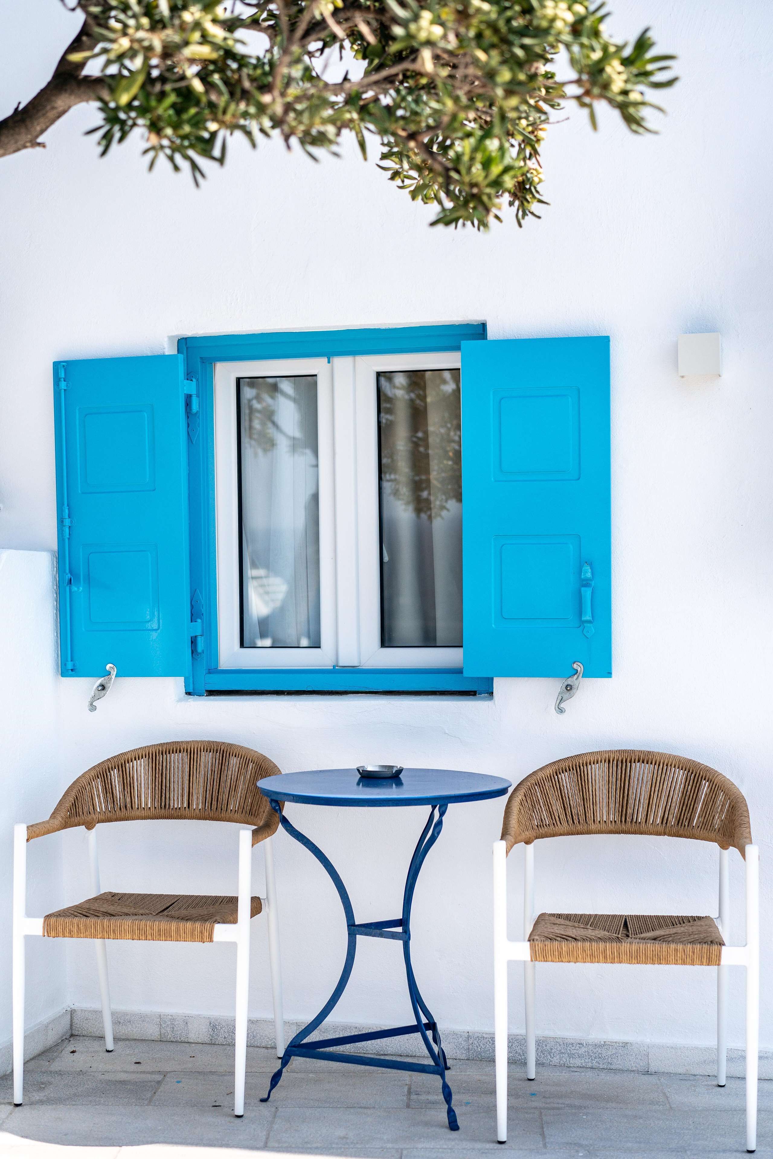 A white wall with a blue window and shutters, two wicker chairs, and a small round table.