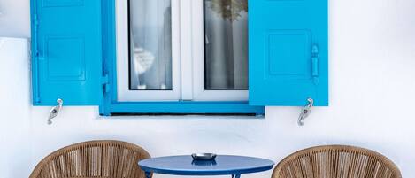 A white wall with a blue window and shutters, two wicker chairs, and a small round table.