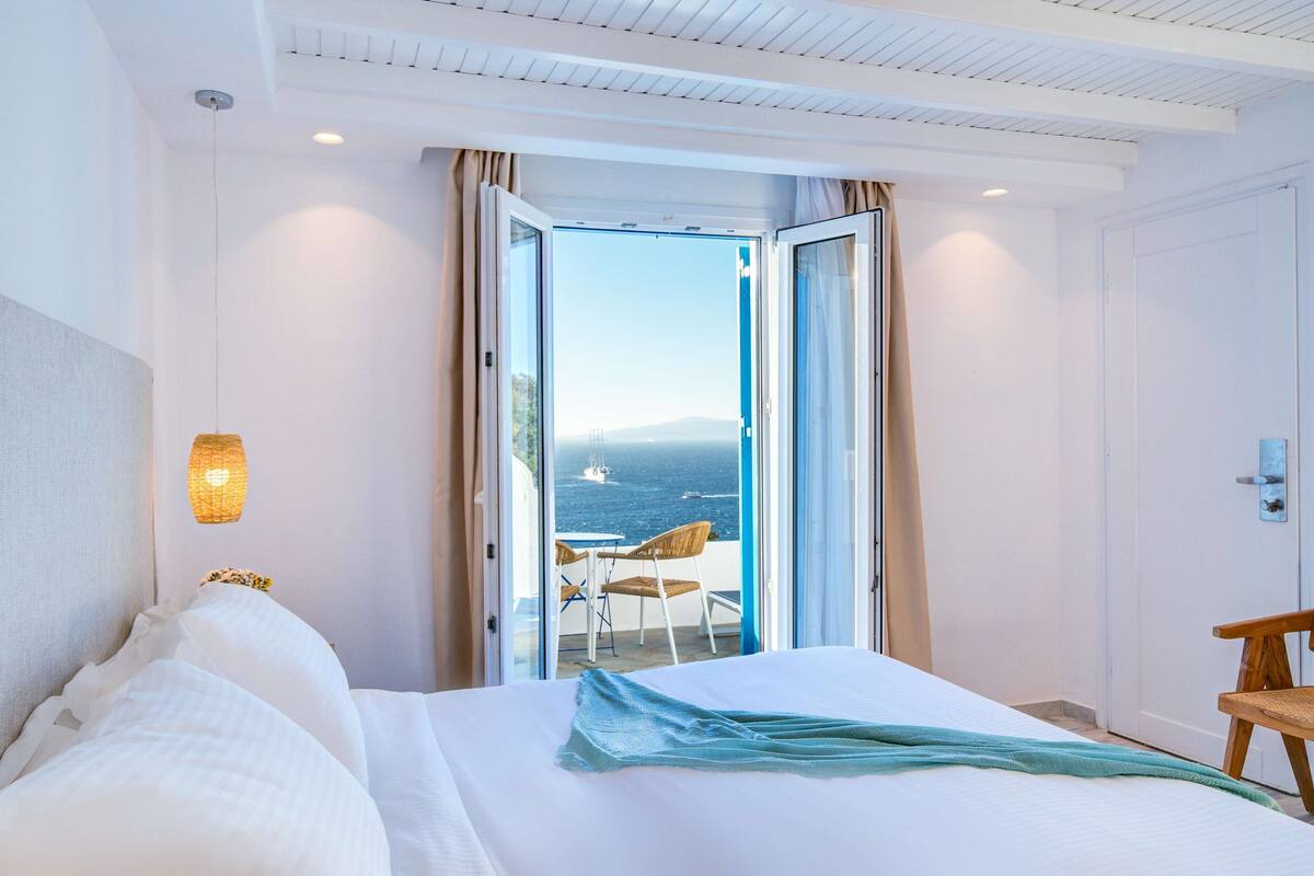 A hotel room with a bed, a chair, a table, and a view of the sea through an open door.
