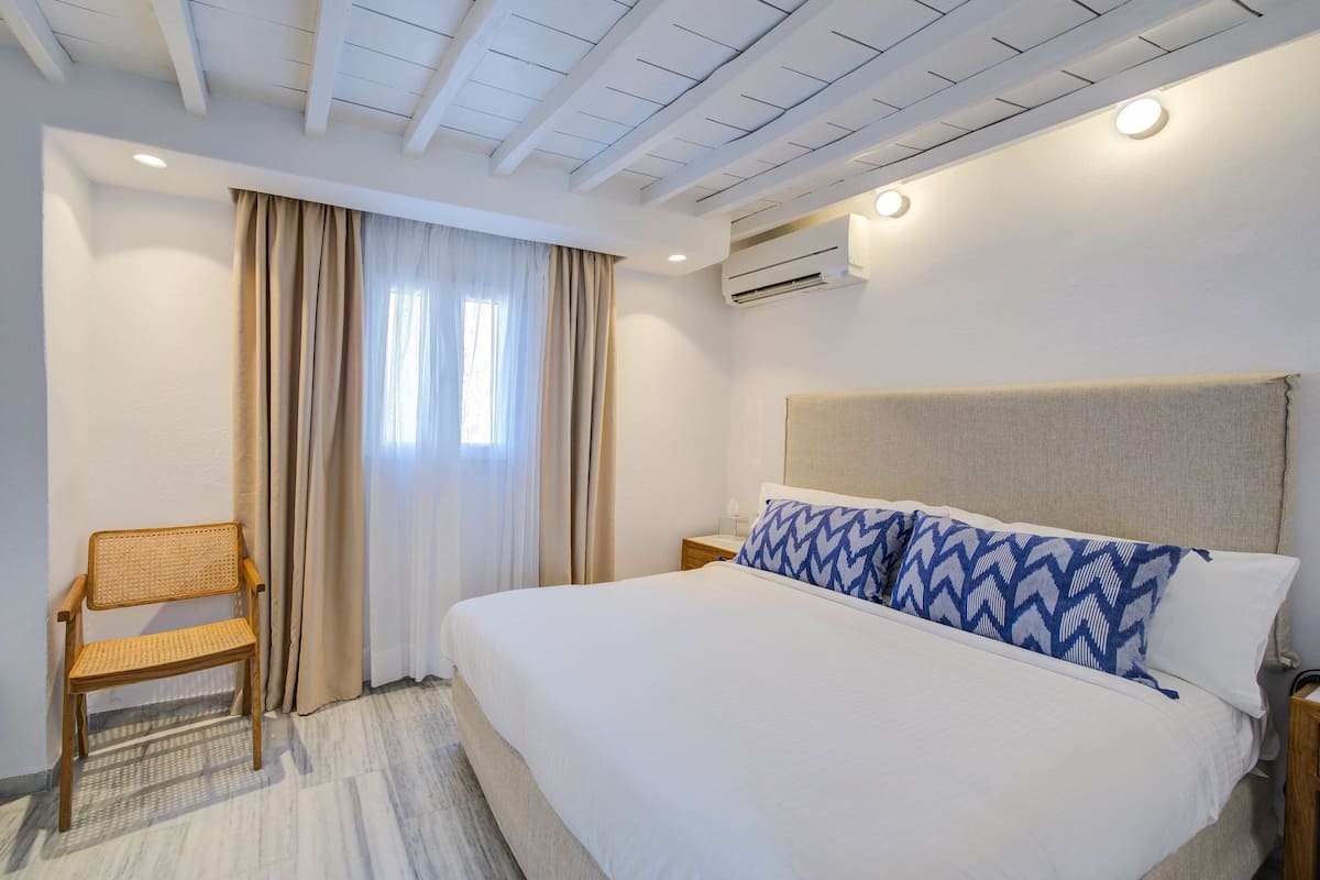 A hotel room with a large bed, a wooden chair, a nightstand, and an air conditioning unit.