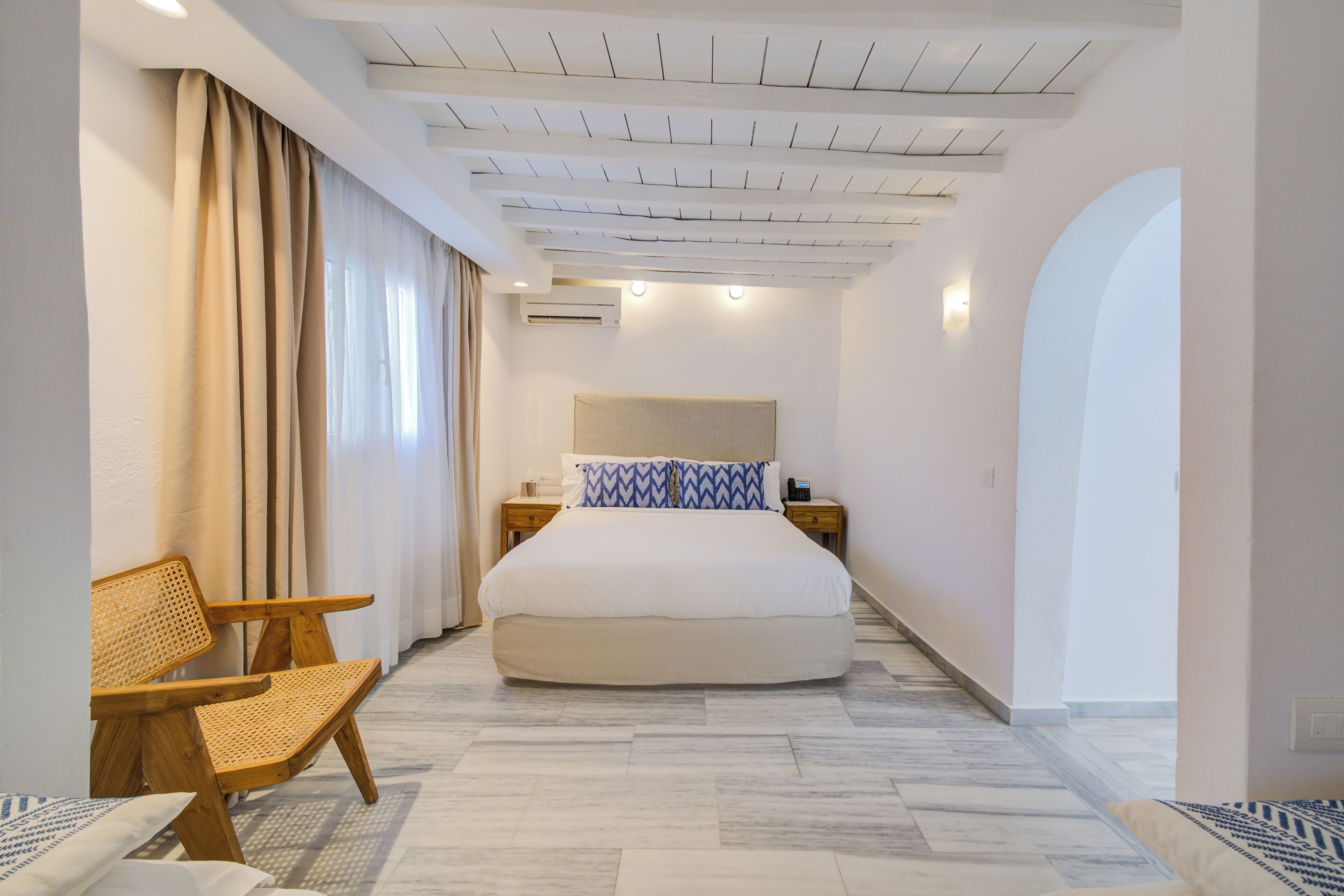 A hotel room with a large bed, a wooden chair, a desk, and a large arched doorway.