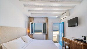 Habitación de hotel con una cama grande, un escritorio, un televisor y vistas al mar a través de ventanas abiertas.