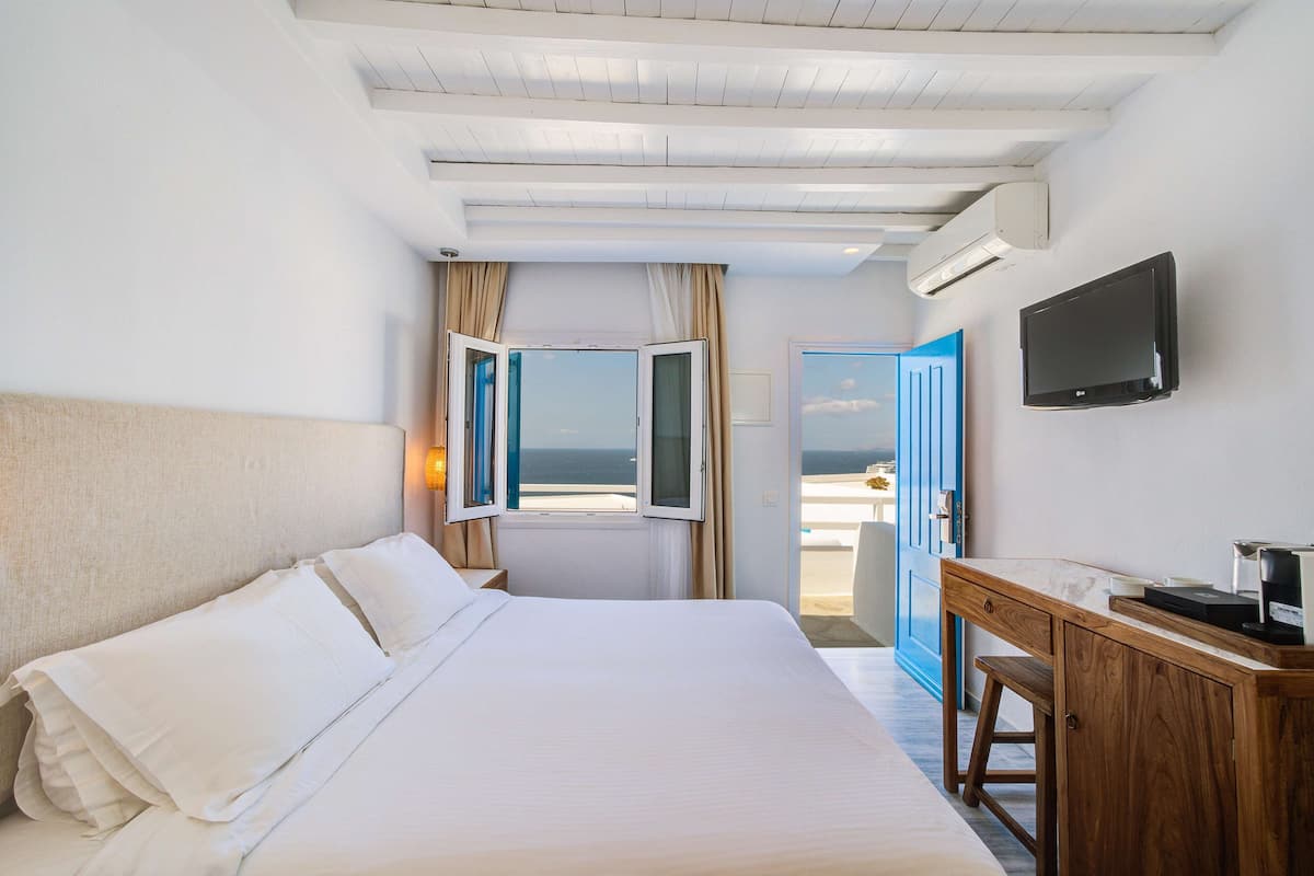 A hotel room with a large bed, a desk, a TV, and a view of the sea through open windows.