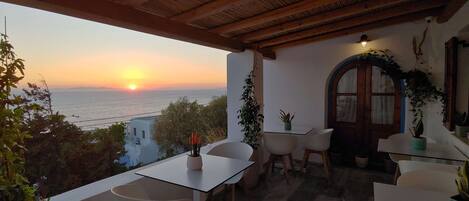 Una terraza con vistas a la puesta de sol sobre el mar, que incluye una mesa y sillas, techo de madera y plantas en macetas.