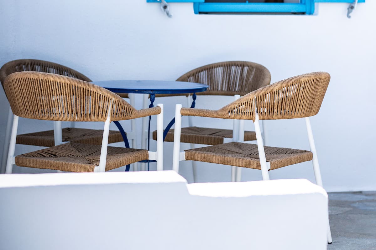 A table with two wicker chairs and a blue tablecloth, set against a white wall with a blue window shutter.
