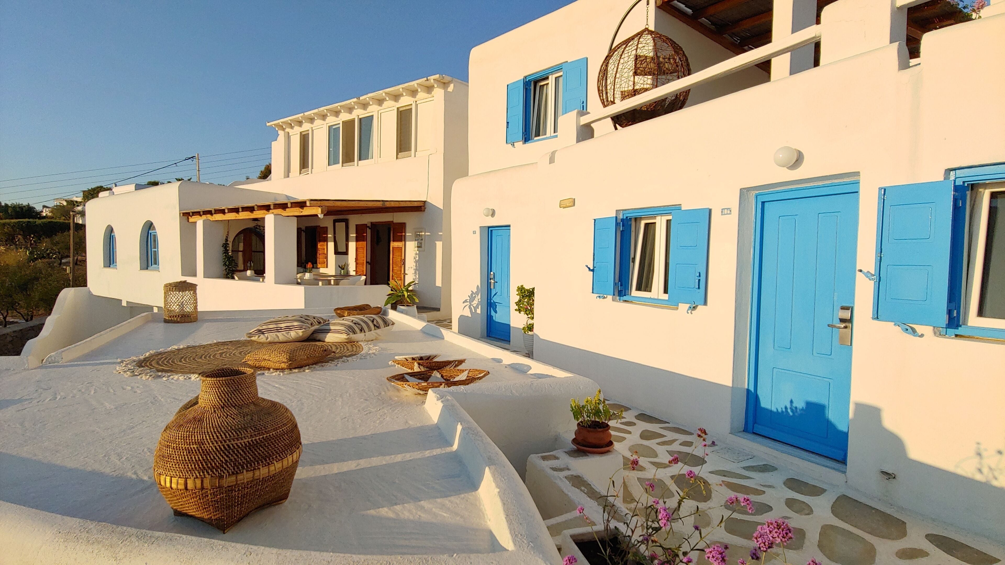 A white building with blue shutters and doors, a covered outdoor seating area with wicker furniture, and a stone-paved patio.