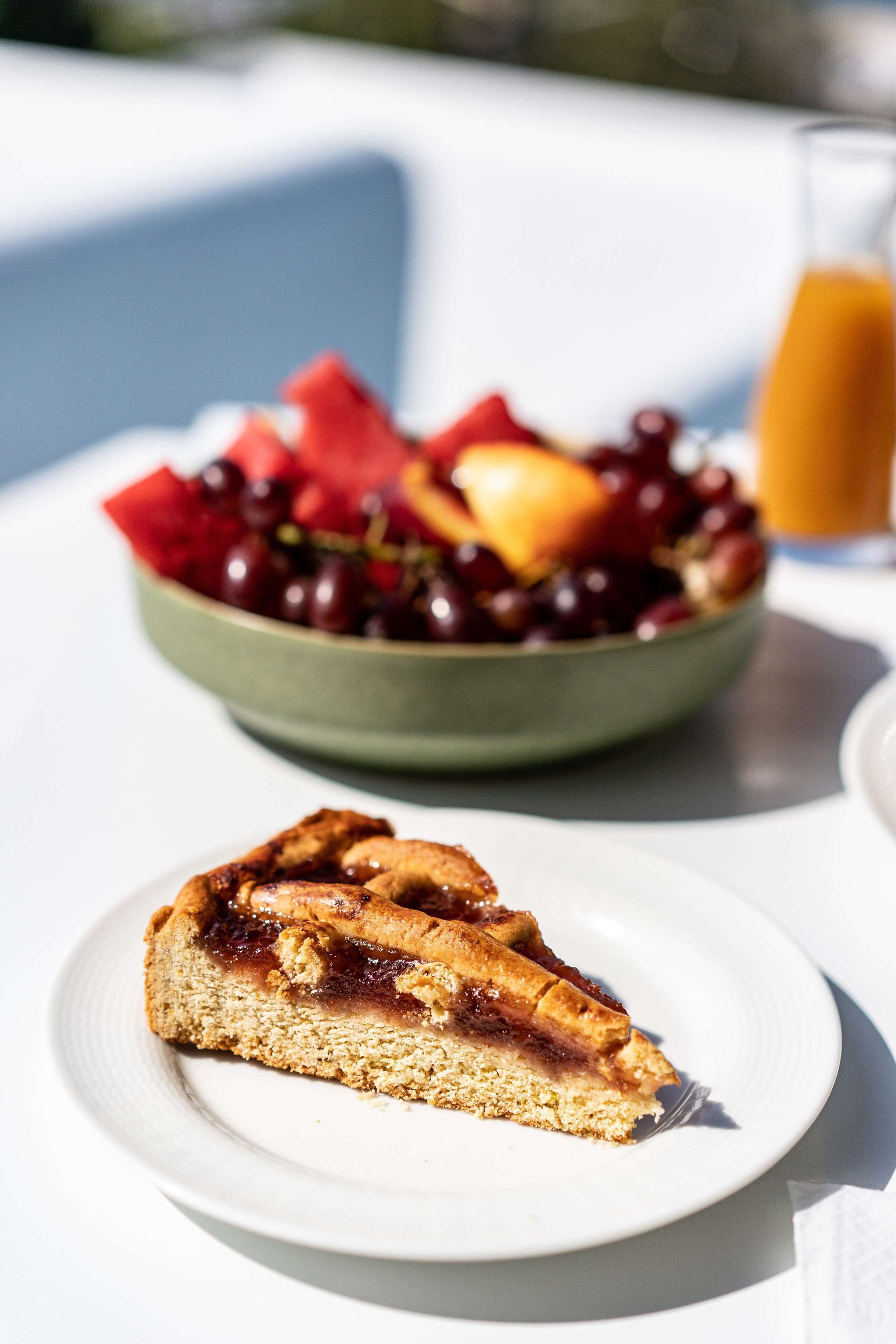 Una porción de pastel en un plato blanco, un bol de frutas variadas y un vaso de zumo de naranja sobre una mesa.