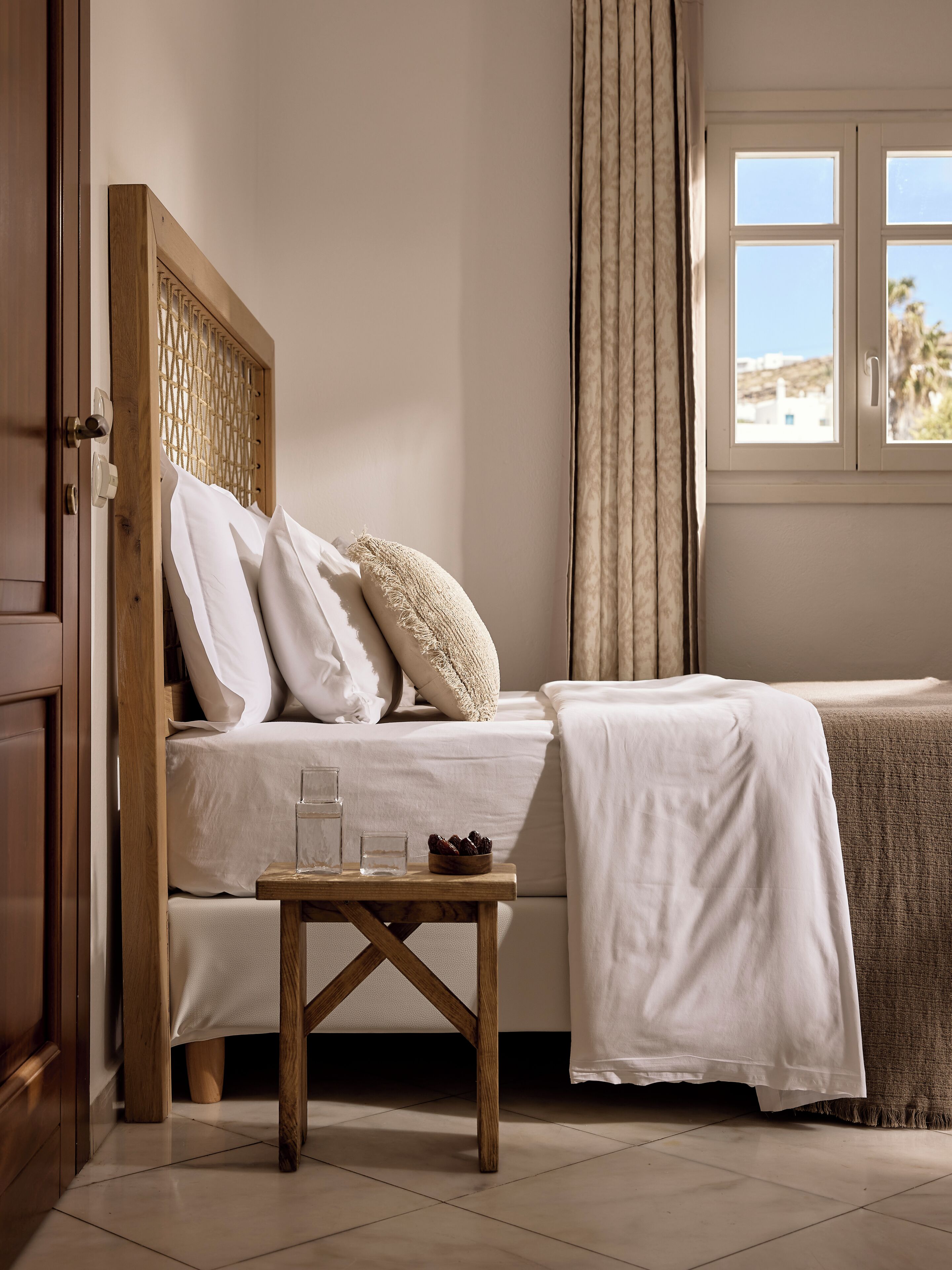 A bedroom with a bed, a wooden headboard, a bedside table with glasses and a bowl of dates, and a window with curtains.