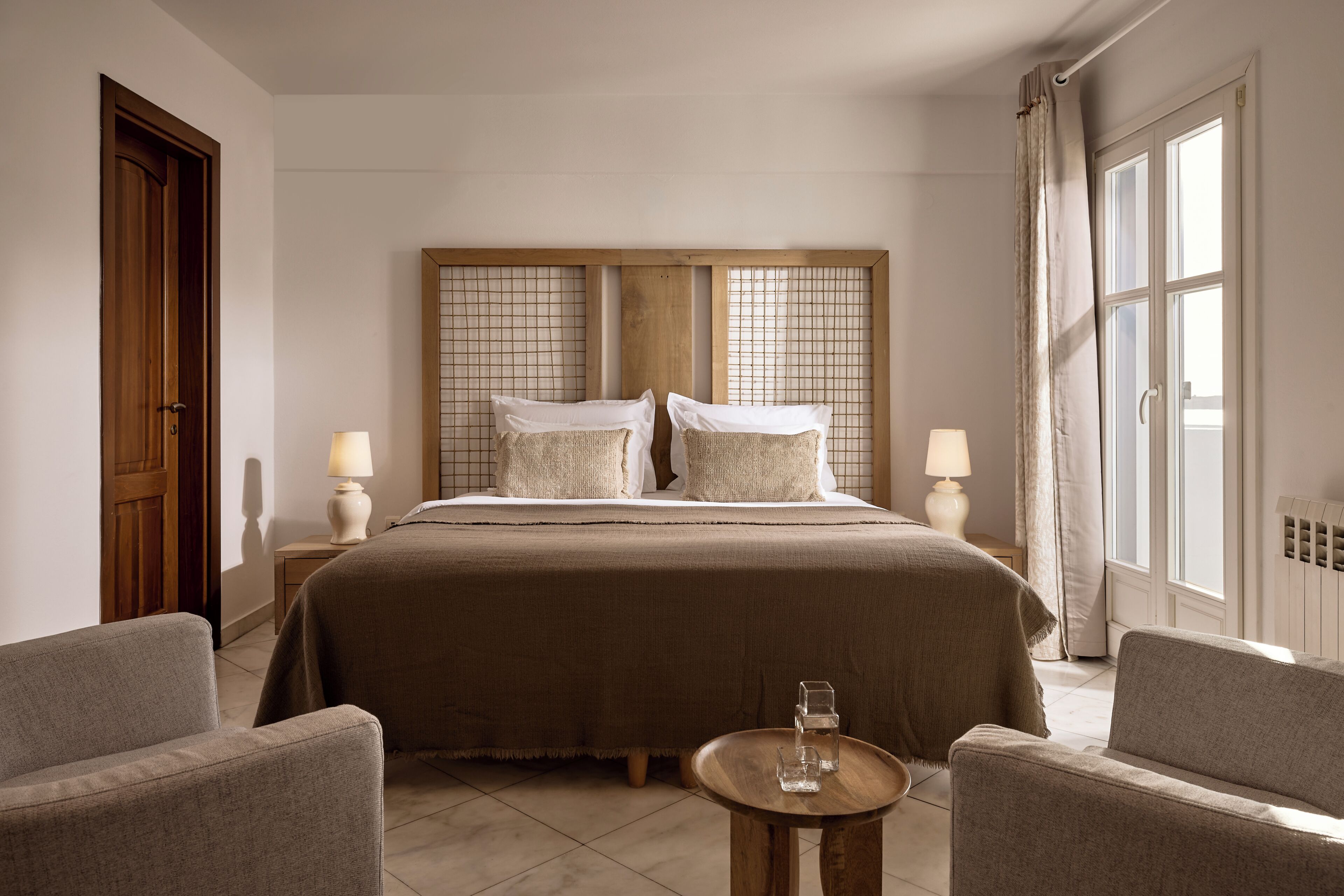 Habitación de hotel con una cama grande, dos mesitas de noche con lámparas, una pequeña mesa redonda con un vaso y dos sillones.