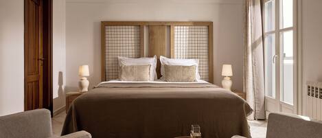 Habitación de hotel con una cama grande, dos mesitas de noche con lámparas, una pequeña mesa redonda con un vaso y dos sillones.