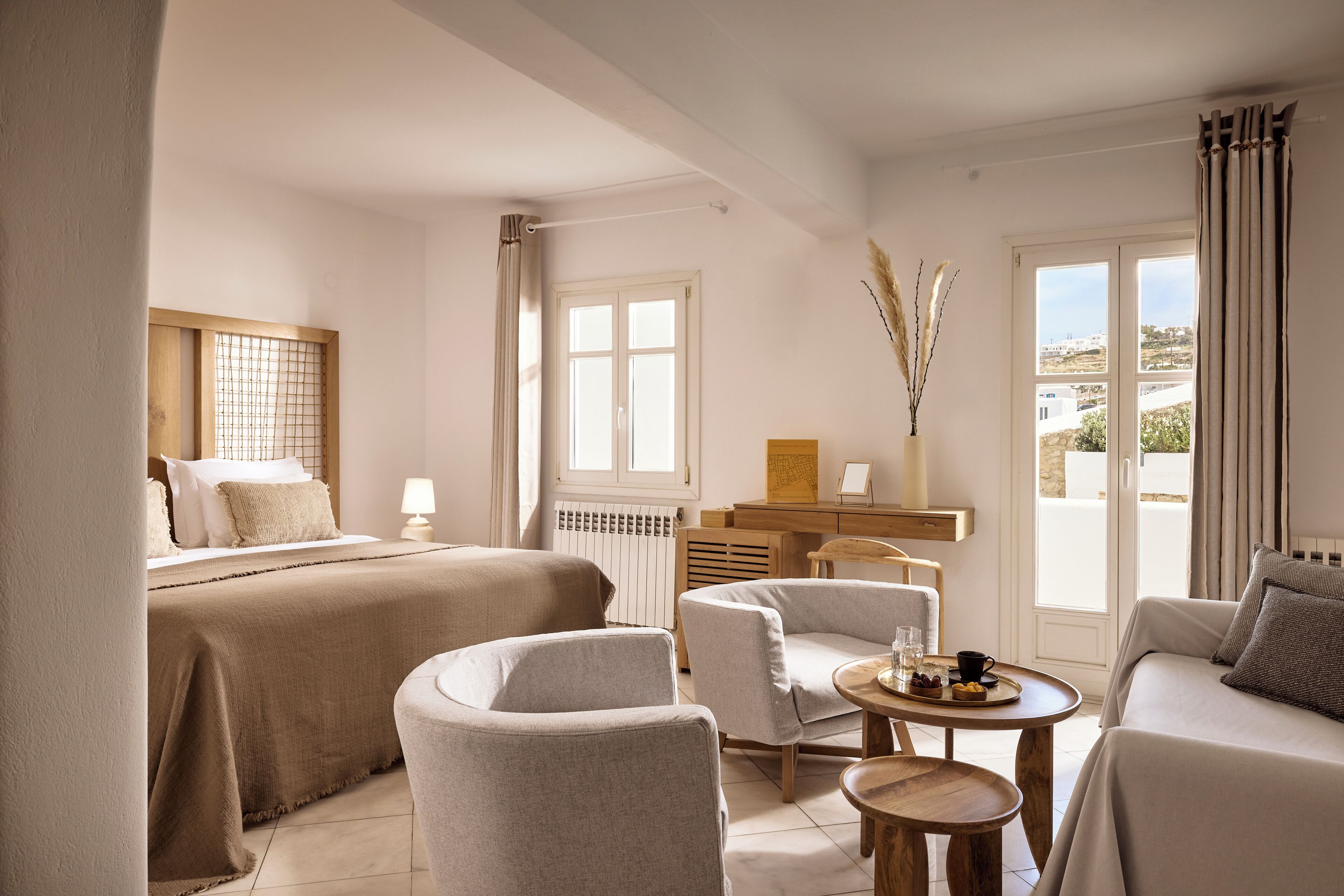 Habitación de hotel con dos camas, un área de estar con mesa y sillas, y vista a la ciudad desde las ventanas.
